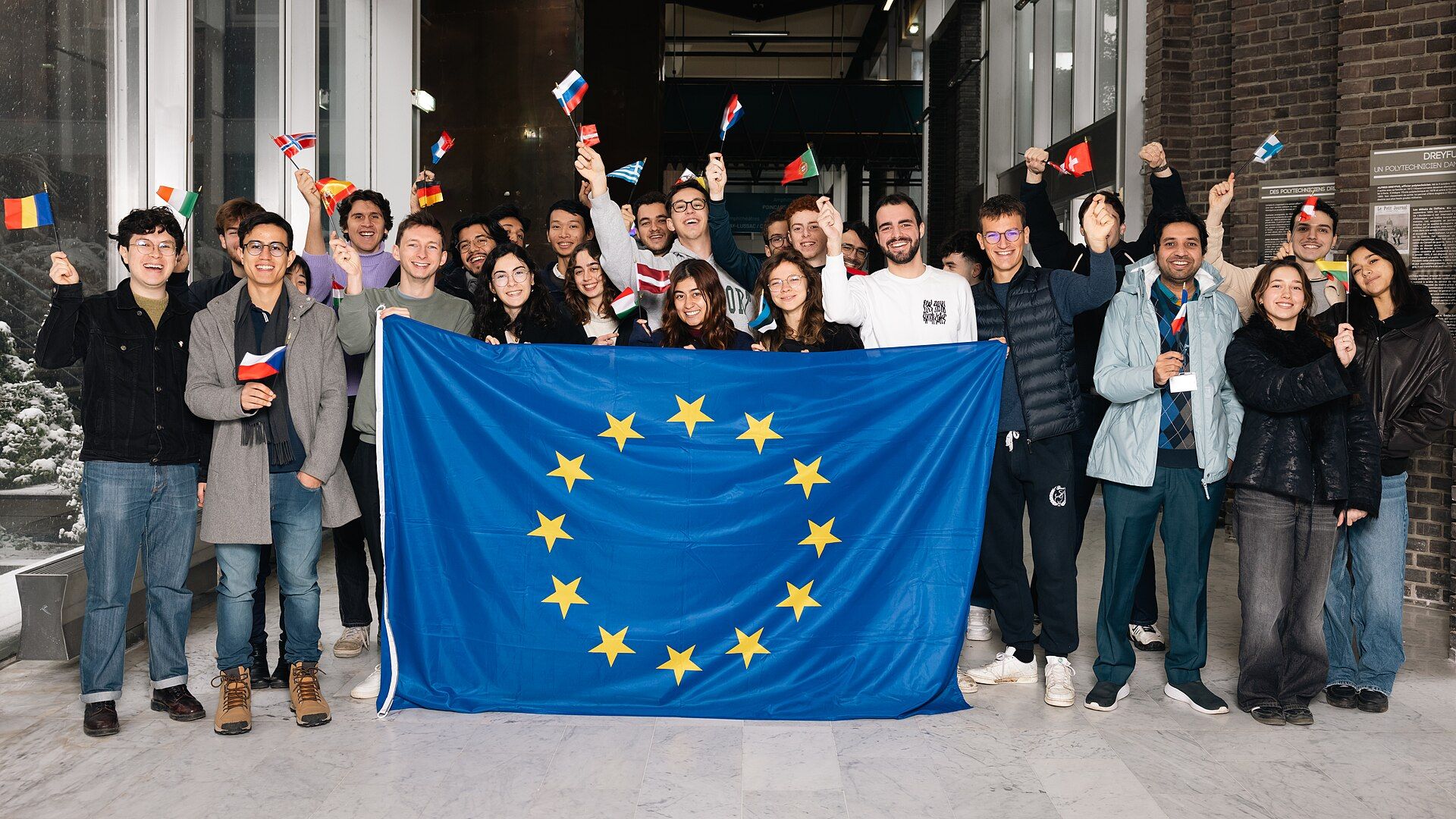 A group of students holding up a large EU flag and small national flags indoors
