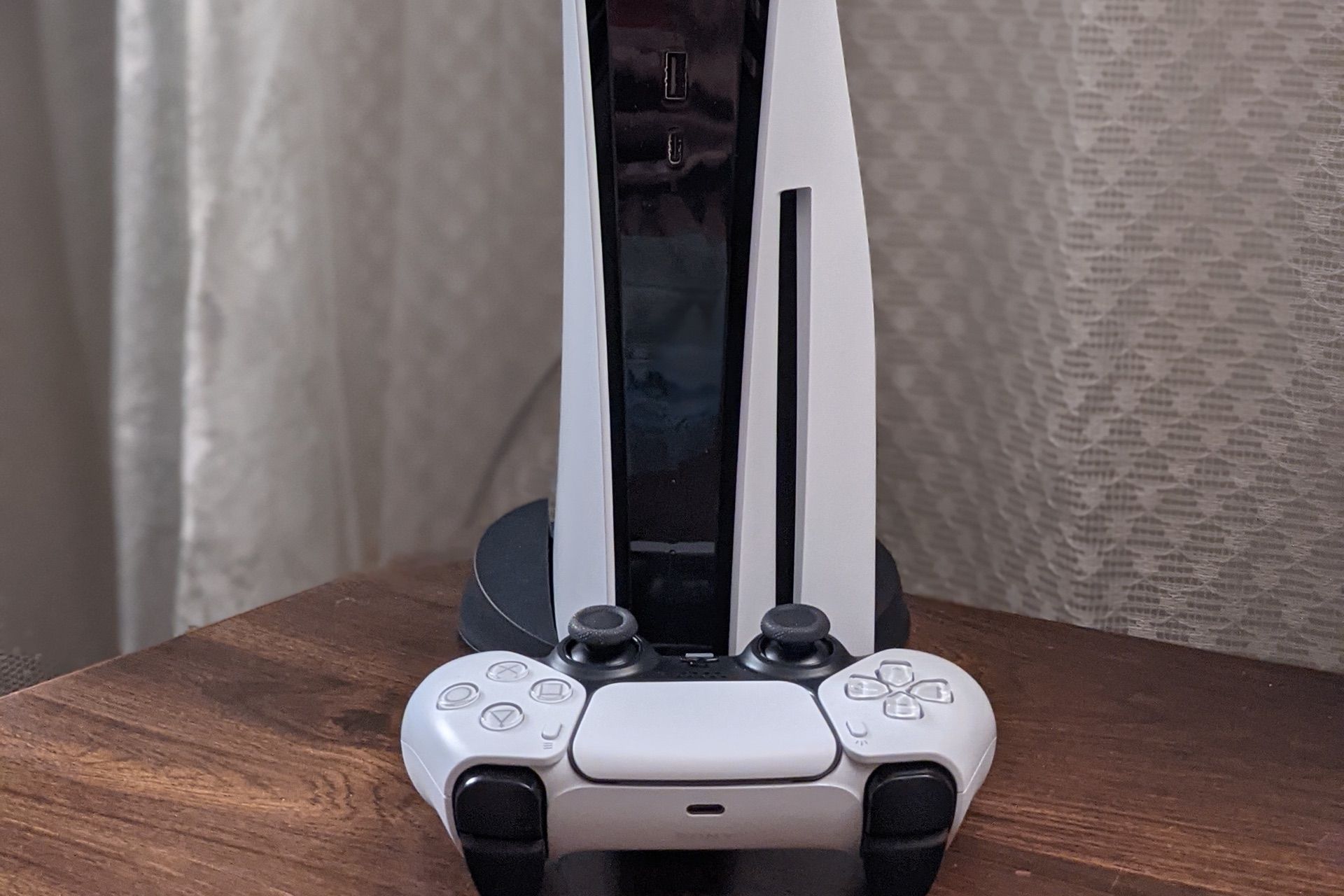 A white PlayStation 5 console standing upright, with its disc slot visible, and a controller in front of it on a wooden table