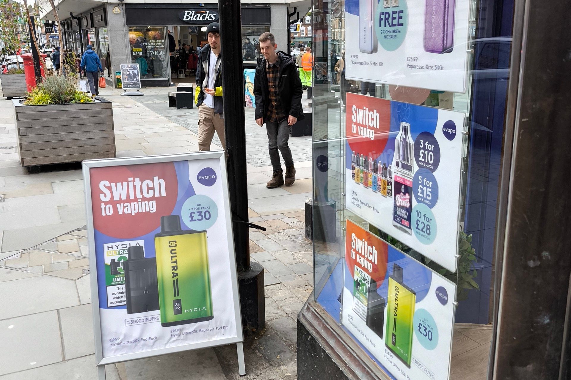 A UK high street vape shop window and A-board advertising vape kits, with offers such as a free item and three for £10, as two people walk past